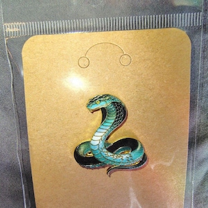 May include: A metal enamel pin shaped like a coiled cobra snake. The snake features a vibrant turquoise and teal gradient body with black accents, a white underbelly, and a gold-toned outline. The pin is attached to a rectangular brown card.