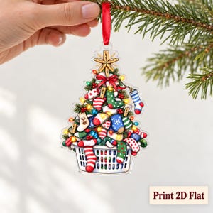 May include: A flat, clear acrylic Christmas tree ornament featuring a graphic illustration of a white laundry basket overflowing with colorful patterned socks, topped with a wooden star and a red ribbon for hanging, with the text Print 2D Flat.