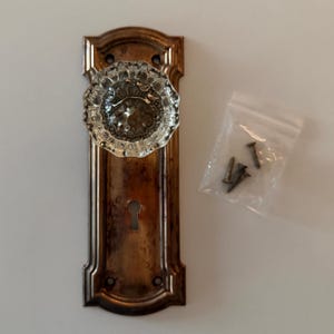 May include: A clear, faceted glass doorknob mounted on a rectangular, weathered bronze-toned metal backplate with a central keyhole. A small, transparent plastic bag containing four metal screws is positioned to the right of the plate.