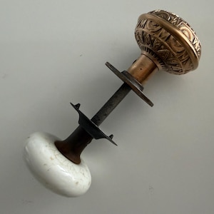 May include: A door knob set featuring one round white ceramic knob with a subtle crackle finish and one round metallic bronze knob with an intricate embossed geometric pattern, connected by a central metal spindle and a flat circular mounting plate.