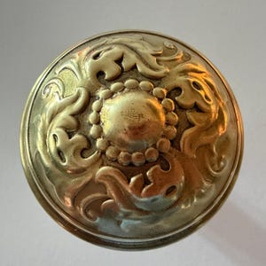 May include: A round brass door knob featuring a central circular dome surrounded by a ring of small beads and an outer border of embossed acanthus leaf scrollwork. The metal surface has a polished gold-toned finish with subtle highlights.