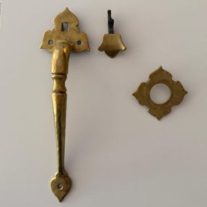 May include: A brass door handle set featuring a long, curved handle with a decorative pointed top and bottom, a separate small metal latch piece, and a square-shaped escutcheon plate with a central circular opening, all with a polished finish.