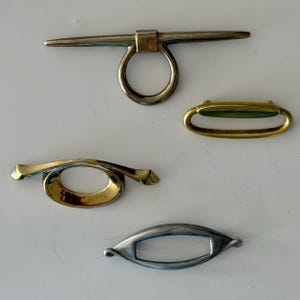 May include: A collection of four metal toggles and fasteners. The top piece is a silver-toned bar with a circular ring. Below it is a gold-toned oval loop, a curved gold-toned fastener with a central opening, and a silver-toned almond-shaped toggle.