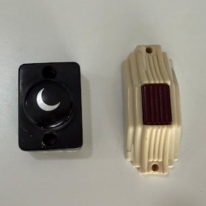 May include: A rectangular black doorbell button featuring a silver crescent moon icon and a cream-colored doorbell button with a dark red rectangular center, both designed for wall mounting with visible screw holes at the top and bottom.
