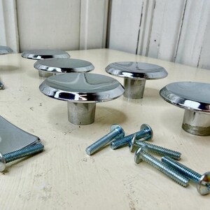 May include: A collection of polished chrome cabinet knobs with a smooth, rounded top and a cylindrical base. The set includes several square metal backplates with a central square hole and a group of threaded metal screws for installation.
