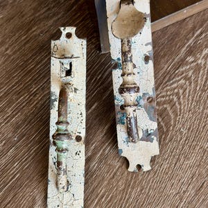 May include: Two vertical metal door handles with rectangular backplates featuring a distressed, cracked off-white paint finish. The handles exhibit visible rust and dark brown metal underneath the chipped paint, with mounting holes at the ends.