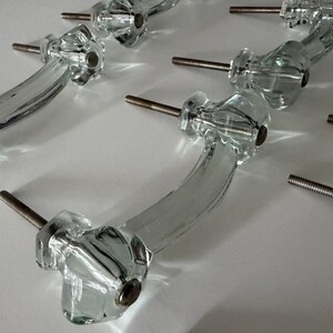 May include: A collection of clear glass drawer pulls featuring a curved handle design with faceted ends. Each pull includes a metallic threaded bolt extending from the base for installation, showcasing a transparent and reflective finish.