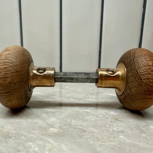 May include: A door knob set featuring two spherical, light brown wood handles connected by a central metal spindle. Each handle is attached to a circular brass-toned metal base, with a visible screw securing the spindle to the brass hardware.