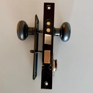 May include: A black mortise door lock set featuring two round dark metal knobs, a rectangular faceplate with a vertical orientation, and a latch mechanism with silver-toned metal components. The hardware includes mounting holes and a spindle.
