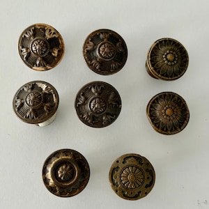 May include: A collection of eight round metal cabinet knobs featuring various embossed floral and geometric patterns in a dark bronze finish. Each knob has a circular face with intricate detailing, including petal motifs and central rosettes.