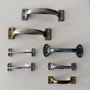 May include: A collection of seven metal drawer pulls and cabinet handles in various finishes, including brushed silver, polished chrome, weathered brass, and matte grey. Each handle features a curved grip and rectangular or oval mounting plates.