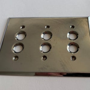 May include: A rectangular, polished chrome wall plate featuring a metallic, reflective finish. The plate includes three circular openings arranged in a vertical column of two, totaling six holes, and three smaller screw holes for mounting purposes.