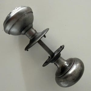 May include: A silver-toned metal door knob set featuring two rounded knobs connected by a square spindle. Each knob is attached to a circular mounting plate with small screw holes, showcasing a weathered, metallic finish and a classic hardware design.