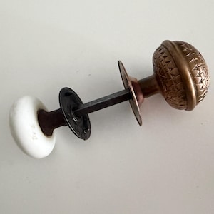 May include: A door knob set featuring one round white porcelain knob and one round brass knob with a textured geometric pattern. The two knobs are connected by a square metal spindle and include a circular black metal mounting plate.
