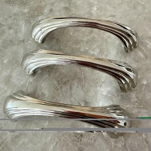 May include: Three polished silver-toned metal drawer pulls arranged vertically. Each handle features a curved, arched shape with a series of parallel, recessed grooves running along the length of the handle, creating a ribbed texture.