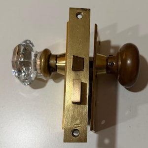 May include: A door hardware set featuring a faceted clear glass knob on one side and a smooth, rounded brass knob on the other. The assembly includes a central brass mortise lock plate with a latch bolt and a deadbolt, connected by a metal spindle.