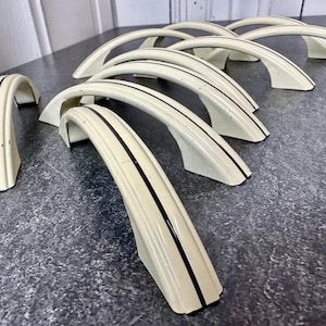 May include: A set of seven arched cabinet pulls in an off-white cream finish, each featuring a prominent black vertical stripe running along the center of the handle. These curved metal hardware pieces are arranged in a staggered formation.