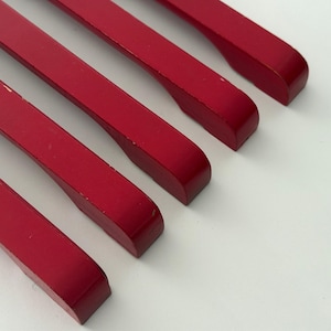 May include: A set of four long, slender, red cabinet pulls arranged diagonally. Each handle features a smooth, matte finish with a slightly curved, ergonomic grip section near the end, showcasing a consistent, vibrant red color throughout.