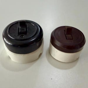 May include: Two round electrical light switches. The larger switch features a black top with a textured center and a white base. The smaller switch has a dark brown top and a white base. Both switches have a central toggle lever for operation.