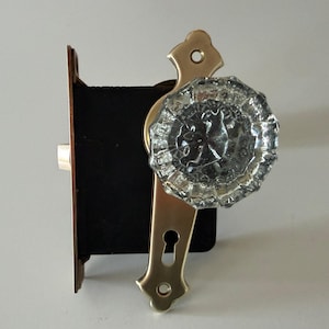 May include: A door knob set featuring a clear, faceted glass knob attached to a polished brass backplate with a keyhole. The mechanism includes a black rectangular mortise lock body with a brass faceplate and a protruding metal latch bolt.