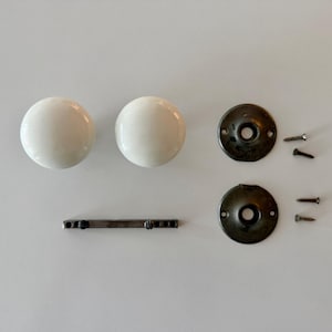 May include: Two round white ceramic door knobs, two circular dark metal mounting plates, a thin rectangular metal bar with two small screws attached, and four additional small metal screws for installation hardware.