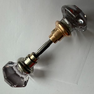 May include: A door knob set featuring two clear, multifaceted glass knobs with an octagonal shape. The knobs are connected by a central metal spindle and include circular brass-toned mounting plates with visible small screws on the side.