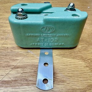 May include: A light green ceramic JFD television lightning arrester, model AT-102, manufactured by J.F.D. Mfg. Co. in Brooklyn, N.Y. The device features two metallic screw terminals on top and a flat, perforated metal mounting bracket extending downward.