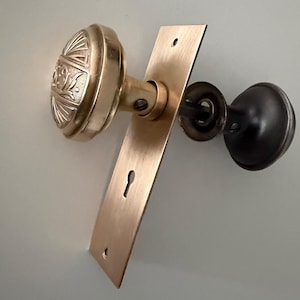 May include: A brass door knob set featuring a round knob with an embossed fan and floral pattern attached to a rectangular brass backplate with a keyhole. The opposite side of the door hardware includes a smaller, plain dark metal round knob.