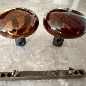 May include: Two round, polished brown and tan marbled door knobs with dark metal bases, each featuring a side hole for attachment. Below the knobs lies a flat, rectangular metal spindle bar with two small screws and multiple circular holes.