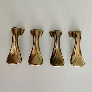 May include: Four metallic furniture feet with a curved, flared design and a brushed gold finish. Each foot features a wide, flat base that tapers upward into a slender neck, topped with a small, angled mounting bracket for attachment to furniture.