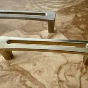 May include: Two metal cabinet pulls with a horizontal rectangular cutout design. One pull features a brushed silver finish, while the other has a polished gold finish. Both hardware pieces have a curved top surface and two sturdy mounting feet.