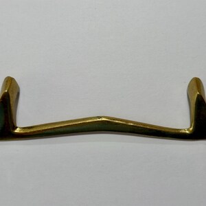 May include: A brass-toned metal drawer pull featuring a wide, arching horizontal bar that curves slightly upward toward the center. The handle has two vertical mounting posts at each end, creating a distinct U-shaped profile with a smooth finish.