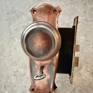 May include: A copper-toned metal door knob set featuring a circular knob mounted on a decorative rectangular backplate with a keyhole. A small metal key is inserted into the keyhole. The mechanism includes a side-mounted latch and strike plate.