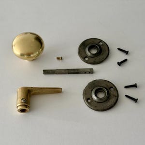 May include: A brass door knob, a brass lever handle, a metal spindle, two circular dark metal mounting plates, and five small black screws. The components are arranged on a plain surface, showing the hardware parts for a door assembly.