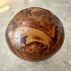 May include: A round cabinet knob featuring a glossy, marbled finish with swirling patterns of dark brown, tan, and cream. The surface displays a subtle network of fine, hairline cracks throughout the glaze, creating a textured appearance.