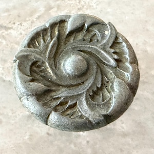 May include: A circular metal knob with a weathered silver-gray finish. The surface features a raised, swirling relief pattern of stylized leaves radiating from a central smooth dome, creating a textured, dimensional appearance on the face.