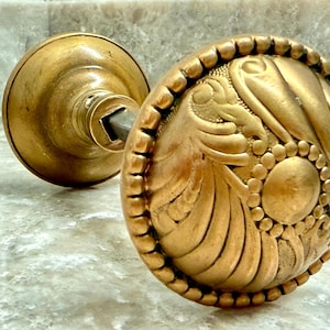 May include: A round brass door knob featuring an intricate embossed pattern of curved, fan-like segments and a central circular medallion. The edge of the knob is decorated with a beaded border, and a square metal spindle extends from the back.