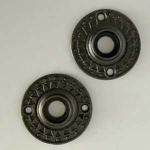 May include: Two circular metal fasteners with a dark gunmetal finish. Each piece features a central hole surrounded by a raised sunburst pattern of triangles and small dots, with two additional small holes positioned on opposite sides of the rim.