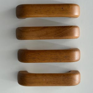 May include: Four rectangular wooden drawer pulls arranged in a vertical column. Each handle features a smooth, rounded profile with a visible natural wood grain pattern in a medium brown tone, showcasing a consistent design across all four pieces.