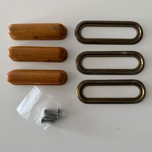 May include: Three light brown wooden drawer pulls with a smooth finish, three matching oval-shaped antique brass metal frames, and a small clear plastic bag containing six silver-colored metal mounting screws for furniture installation.