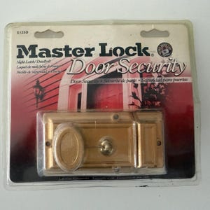 May include: A gold-colored Master Lock door security night latch and deadbolt in its original clear plastic packaging. The packaging features the text Master Lock Door Security and includes a graphic of a red door and a wall-mounted light fixture.