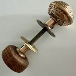 May include: A door knob set featuring one metallic brass knob with an intricate embossed geometric pattern and one smooth, dark brown wooden knob. The two knobs are connected by a central metal spindle and include two circular brass plates.