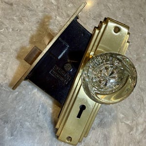 May include: A vintage mortise lock set featuring a clear, faceted glass doorknob attached to a rectangular brass backplate with a keyhole. The internal mechanism is a black metal box with the embossed text SARGENT MADE IN USA visible.