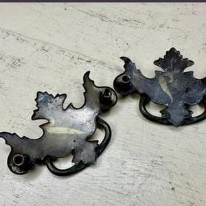 May include: Two dark metal drawer pulls featuring ornate, symmetrical backplates with intricate scrollwork edges and curved, swinging handles. Each backplate includes two circular mounting points on the sides for attachment to furniture surfaces.