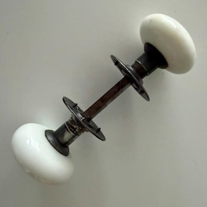 May include: A door knob set featuring two smooth, round, white porcelain knobs connected by a central metal spindle. Each knob is attached to a circular metal mounting plate with small protruding spikes, finished in a dark, weathered metal tone.