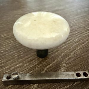 May include: A round, off-white, smooth porcelain door knob with a glossy finish attached to a dark metal base. Below the knob lies a rectangular, flat, metallic spindle bar featuring a single screw head and two circular holes at one end.
