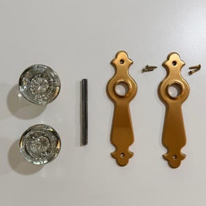 May include: A set of door hardware including two round clear glass knobs with a faceted starburst pattern, one threaded metal spindle, two elongated gold-toned metal backplates with a decorative shape, and four small matching metal screws.