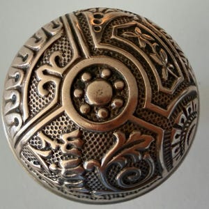May include: A round, metallic door knob featuring an intricate, embossed pattern. The design includes a central circular element surrounded by six small dots, with textured, grid-like sections and raised, swirling floral-inspired motifs.