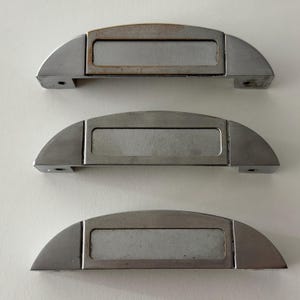May include: Three semi-circular metal drawer pulls with a brushed silver finish. Each handle features a central rectangular label holder frame. The top pull shows slight oxidation around the edges of the frame, while the others are clean.