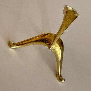 May include: A gold-colored metal display stand featuring a tripod base with three curved legs and a central vertical support arm. The stand has a smooth, reflective finish and is designed to hold objects in an upright position for display.